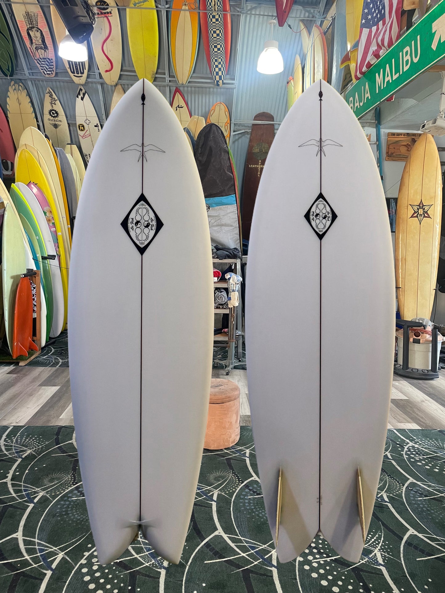 Two white surfboards with a black diamond logo on a patterned floor, surrounded by various other surfboards.