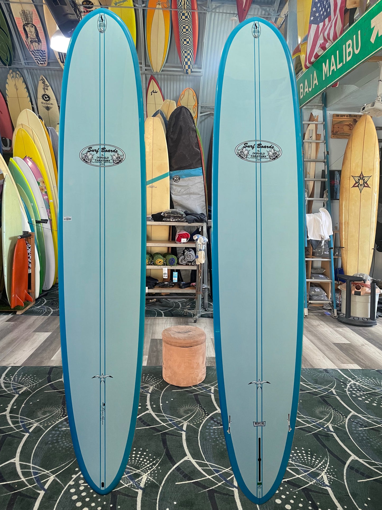 Two blue surfboards side by side in a surf shop with various surfboards and equipment in the background.