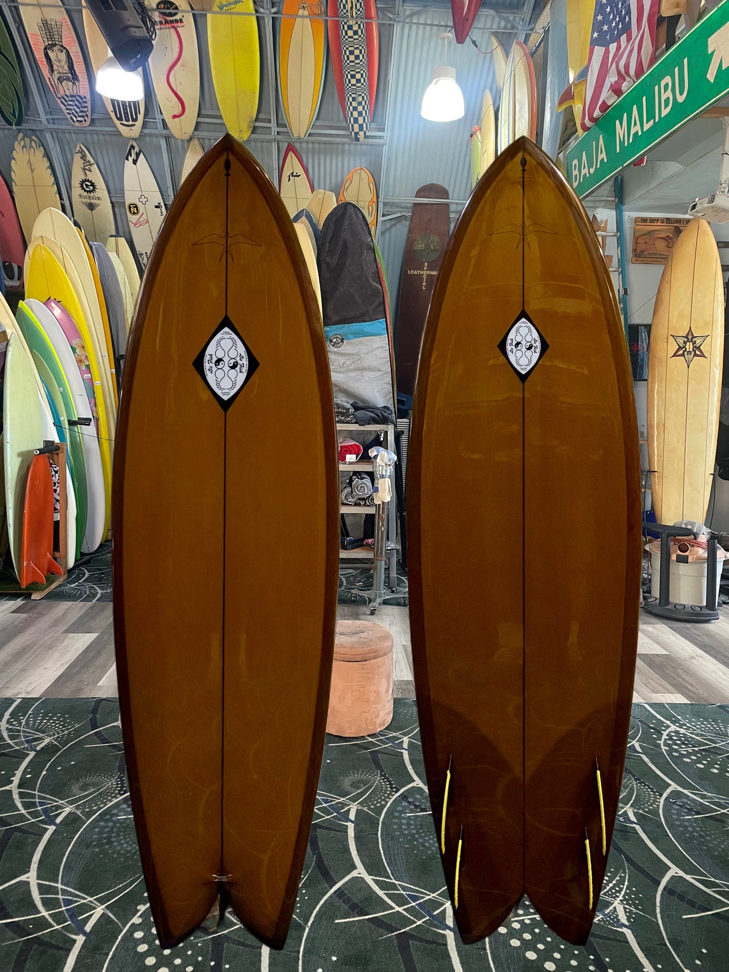 Two brown surfboards with a logo on a patterned floor with other surfboards and decor in the background.