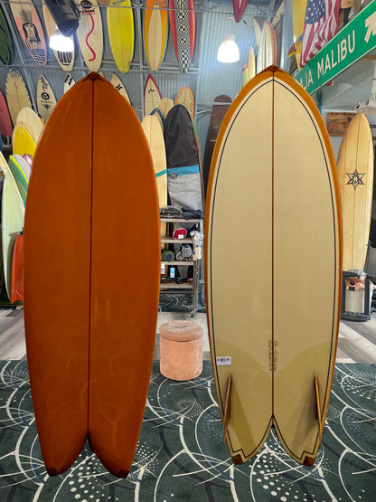 Two surfboards, one orange and one beige, displayed indoors with a patterned floor and various surfboards in the background.