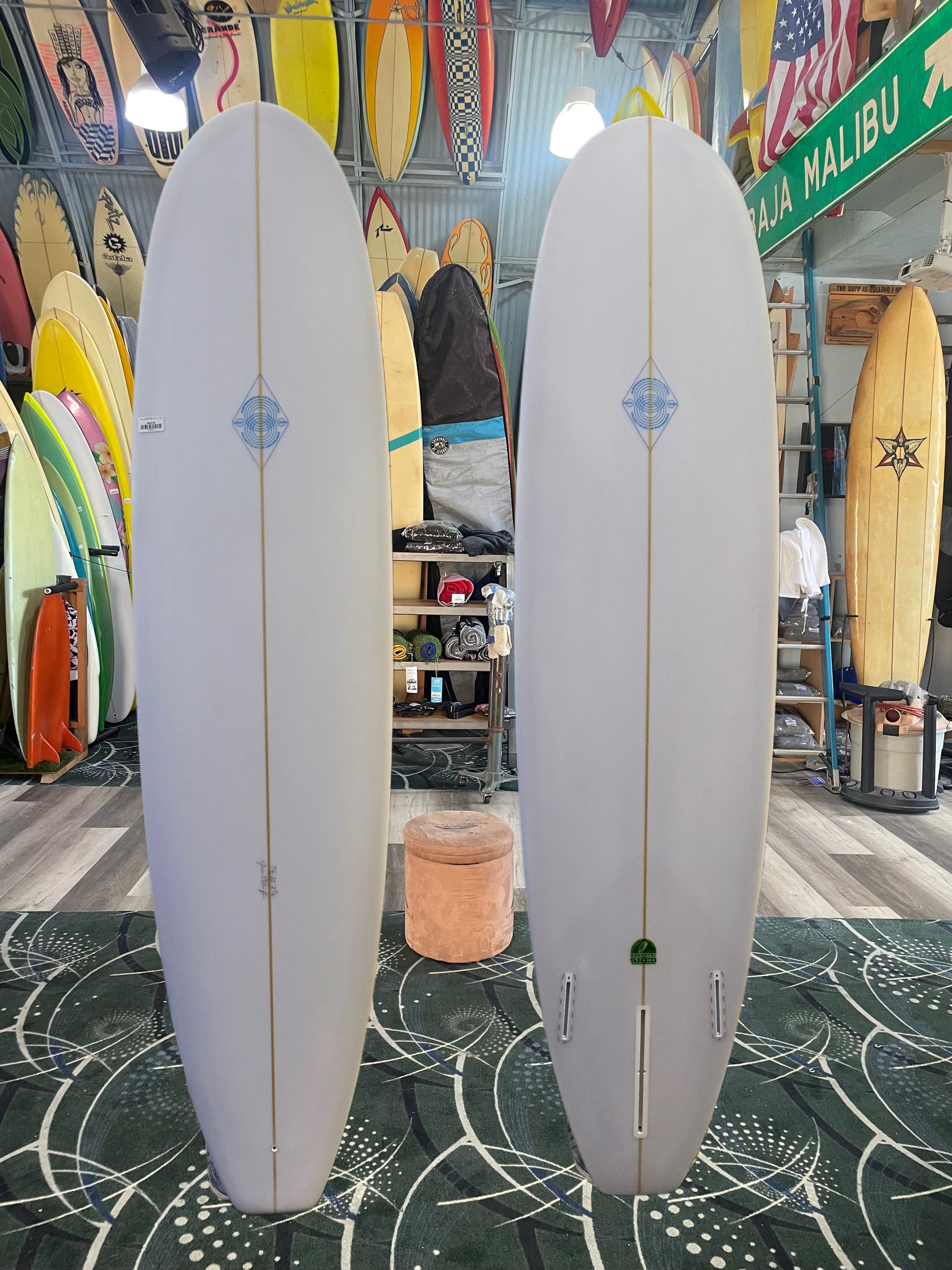 Two white surfboards with blue logos on a green patterned floor in a surf shop.