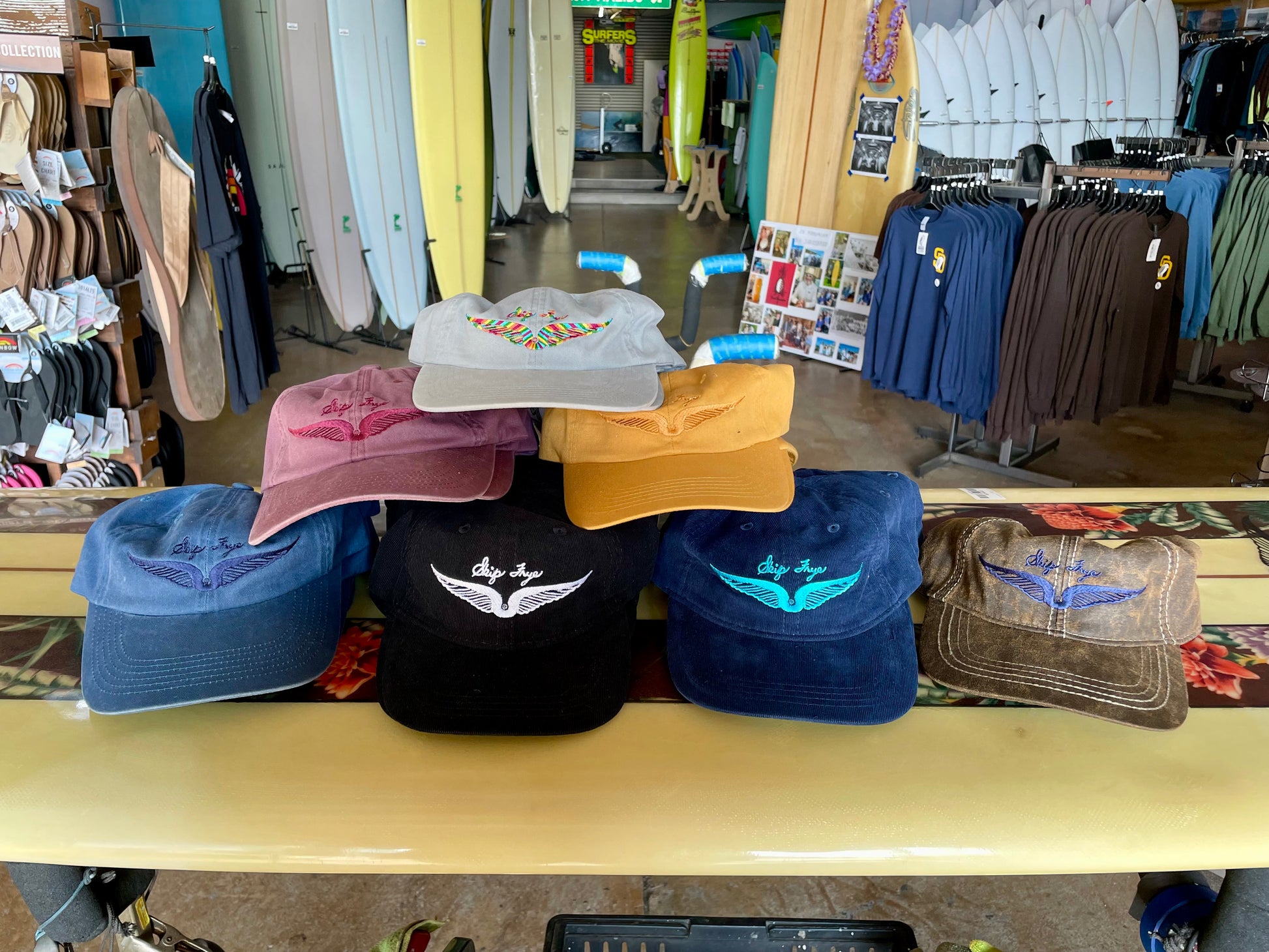 Collection of hats on a table in a store with surfboards and clothing in the background