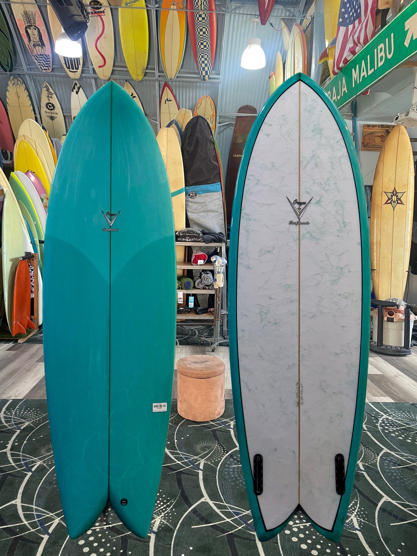 Two surfboards, one teal and one white with black accents, displayed in a shop with other surfboards in the background.