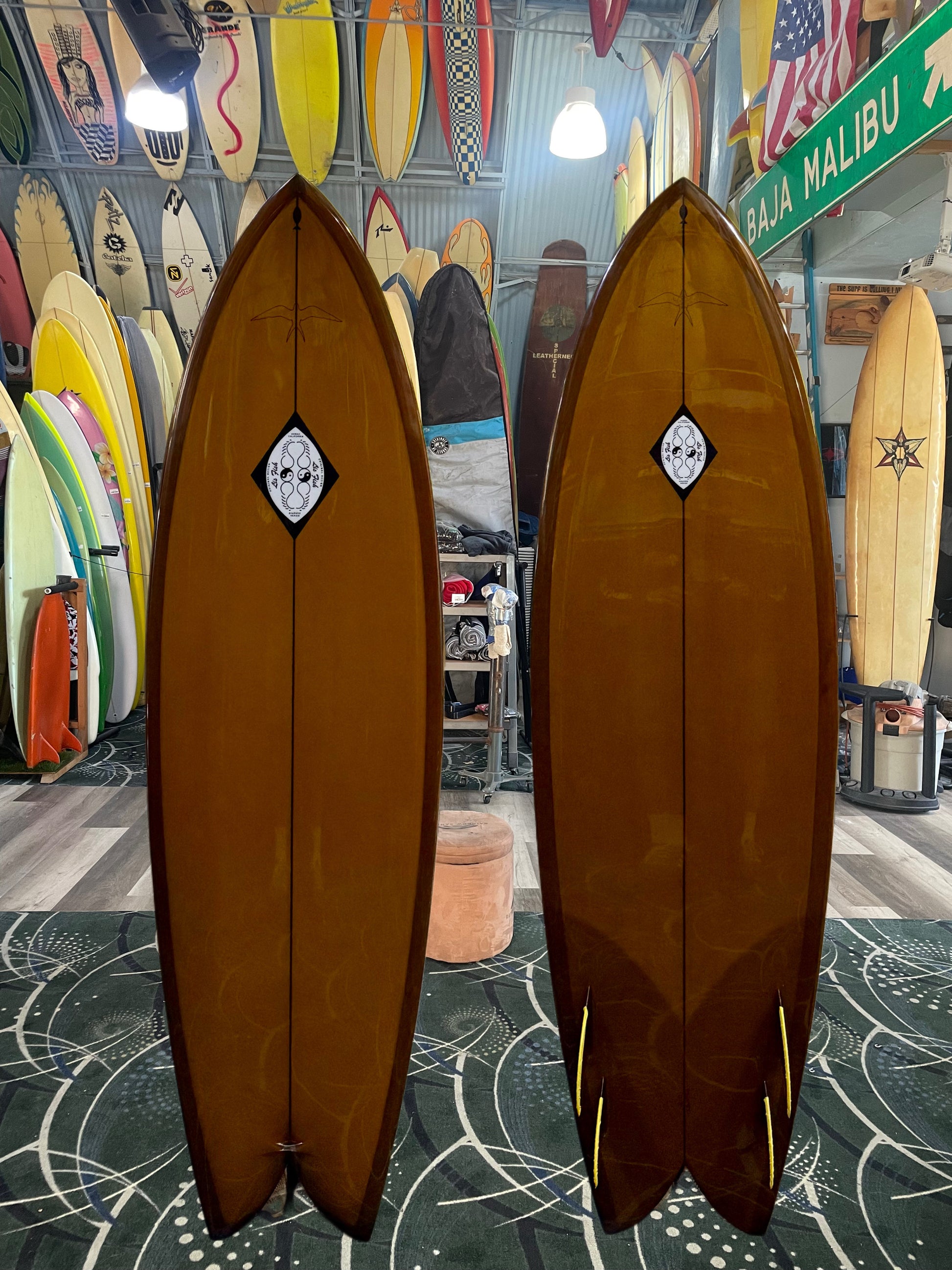 Two brown surfboards with a logo on a patterned floor with other surfboards and decor in the background.