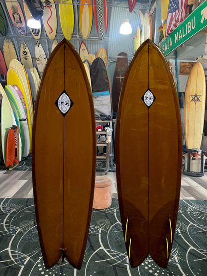 Two brown surfboards with a logo on a patterned floor with other surfboards and decor in the background.