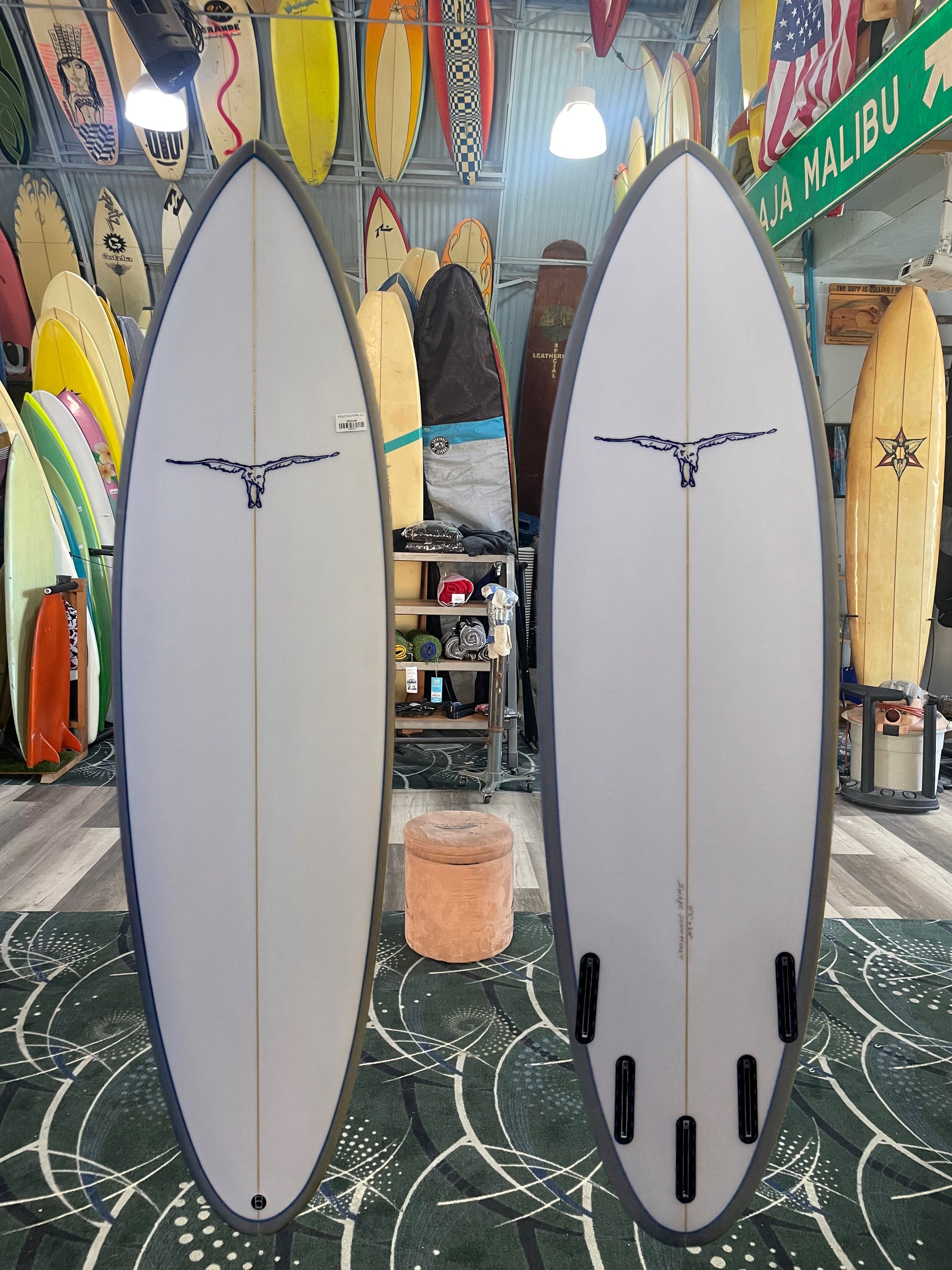 Two surfboards on display in a shop with various surfboards and a 'La Malibu' sign in the background.
