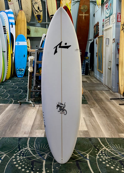White surfboard with black logos in a store setting with other surfboards.