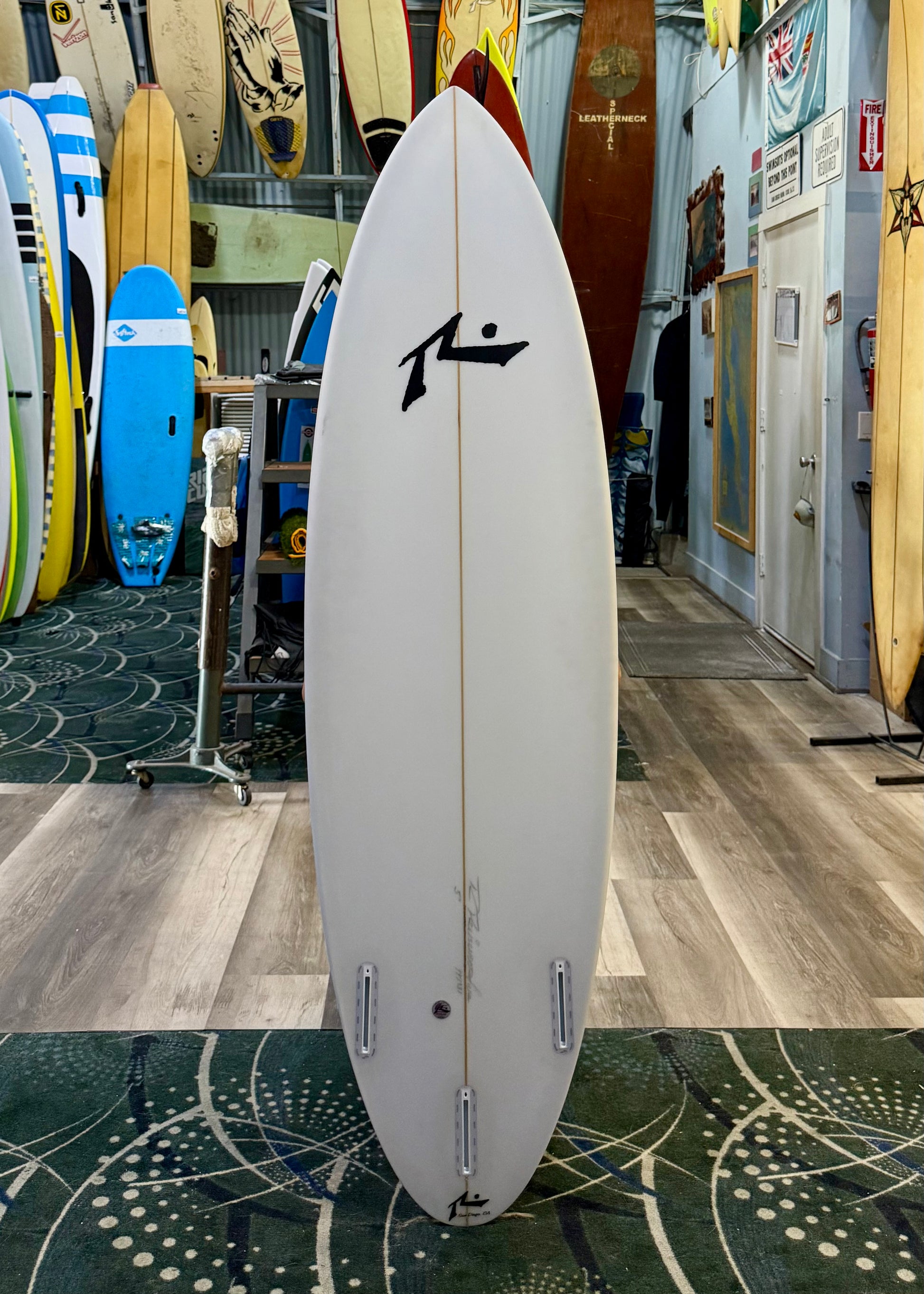 White surfboard with a black logo in a store setting with other surfboards.