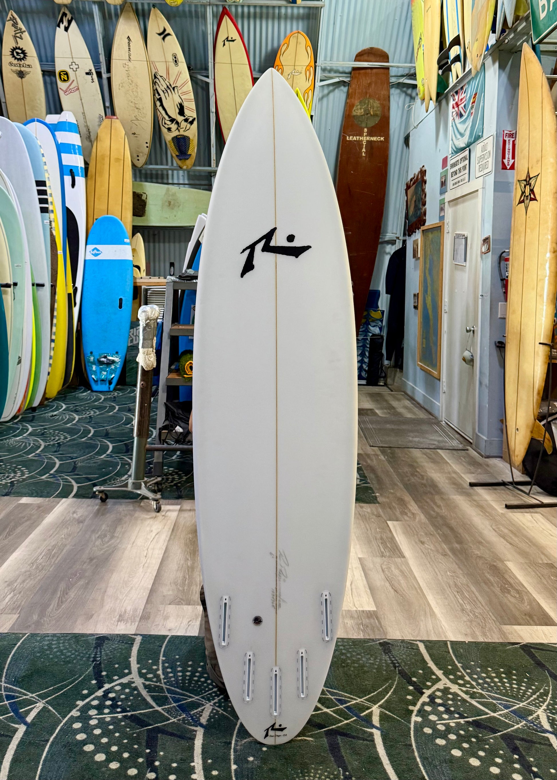 White surfboard with a logo in a surf shop with various other surfboards in the background.