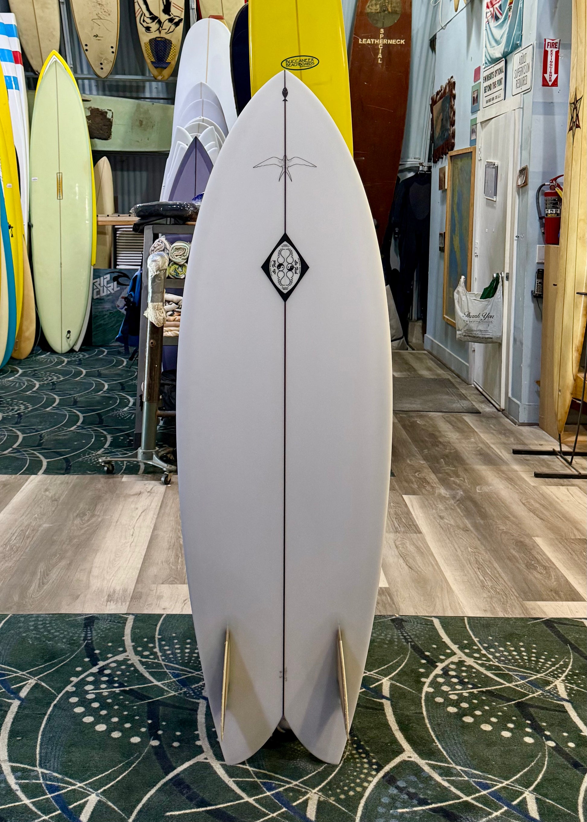 White surfboard with a black diamond logo in a store setting with other surfboards.
