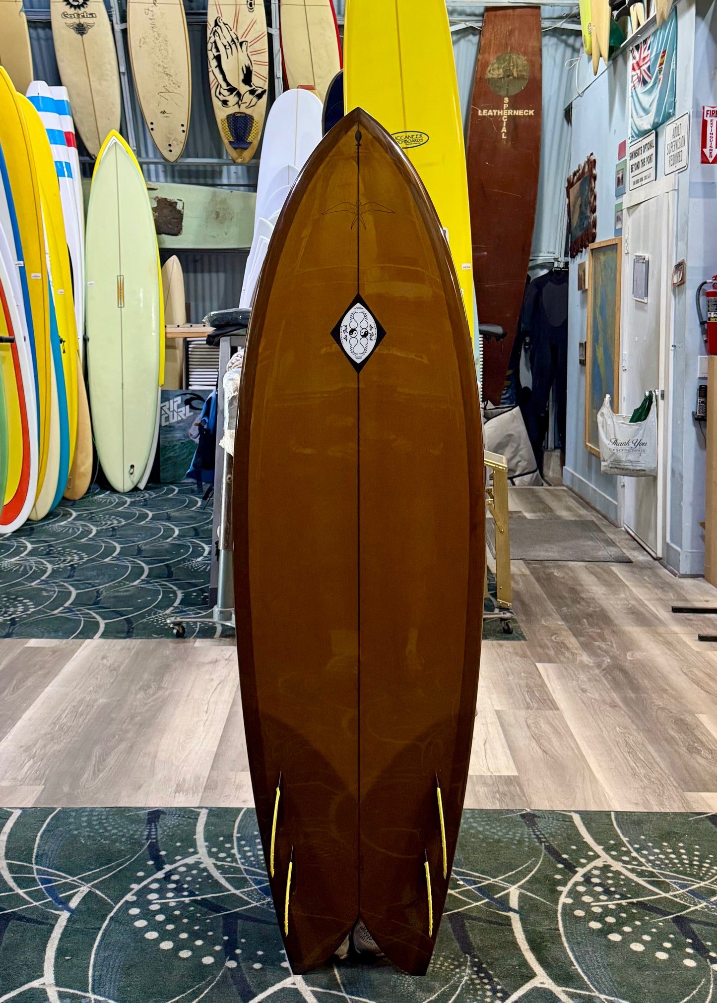 Brown surfboard with a logo on a patterned floor with other surfboards in the background
