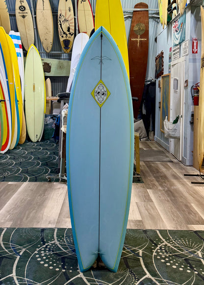 Blue surfboard with a logo on display in a surf shop with other boards in the background.