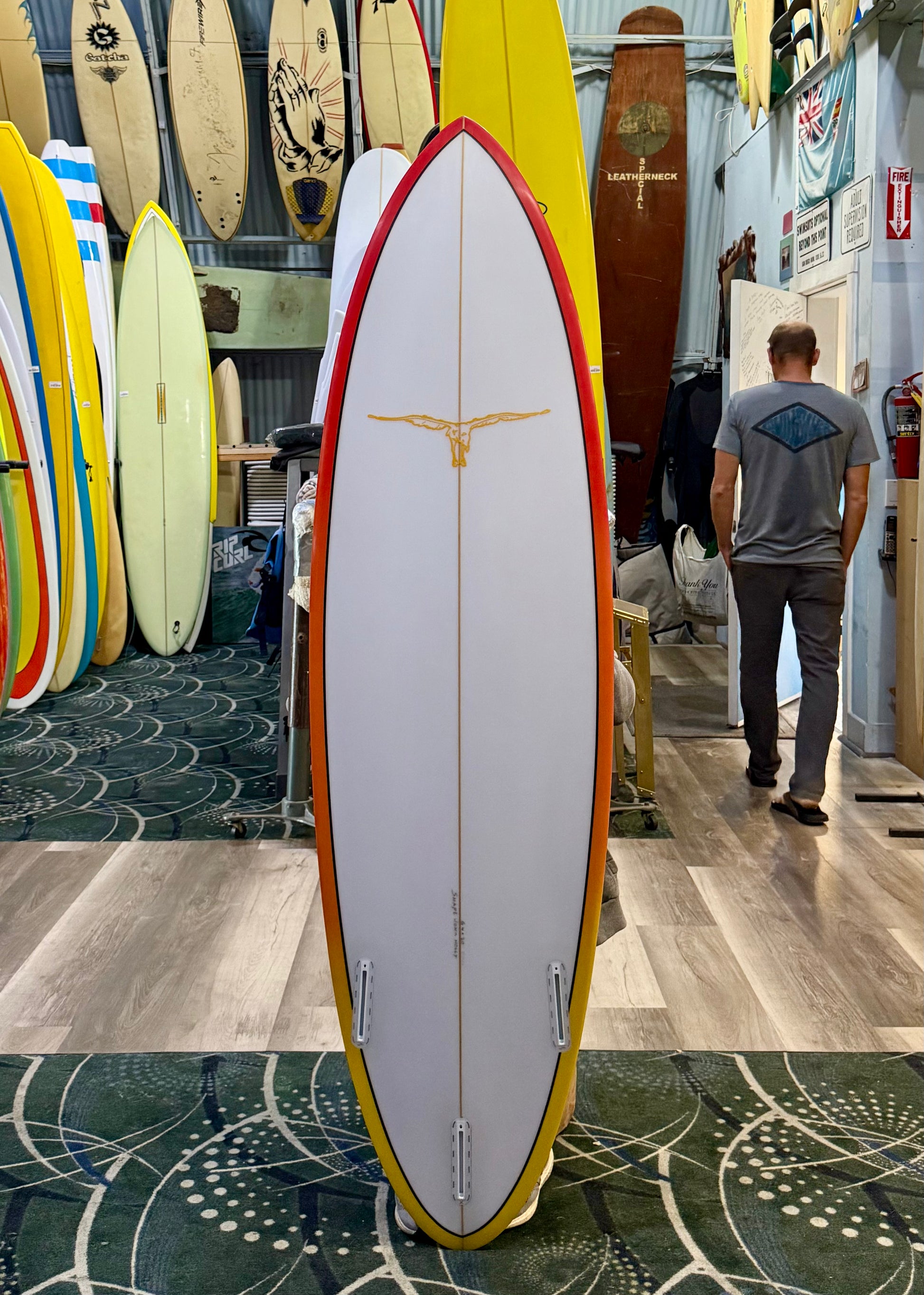 Surfboard with a red and yellow design in a surf shop with other boards and a person in the background.