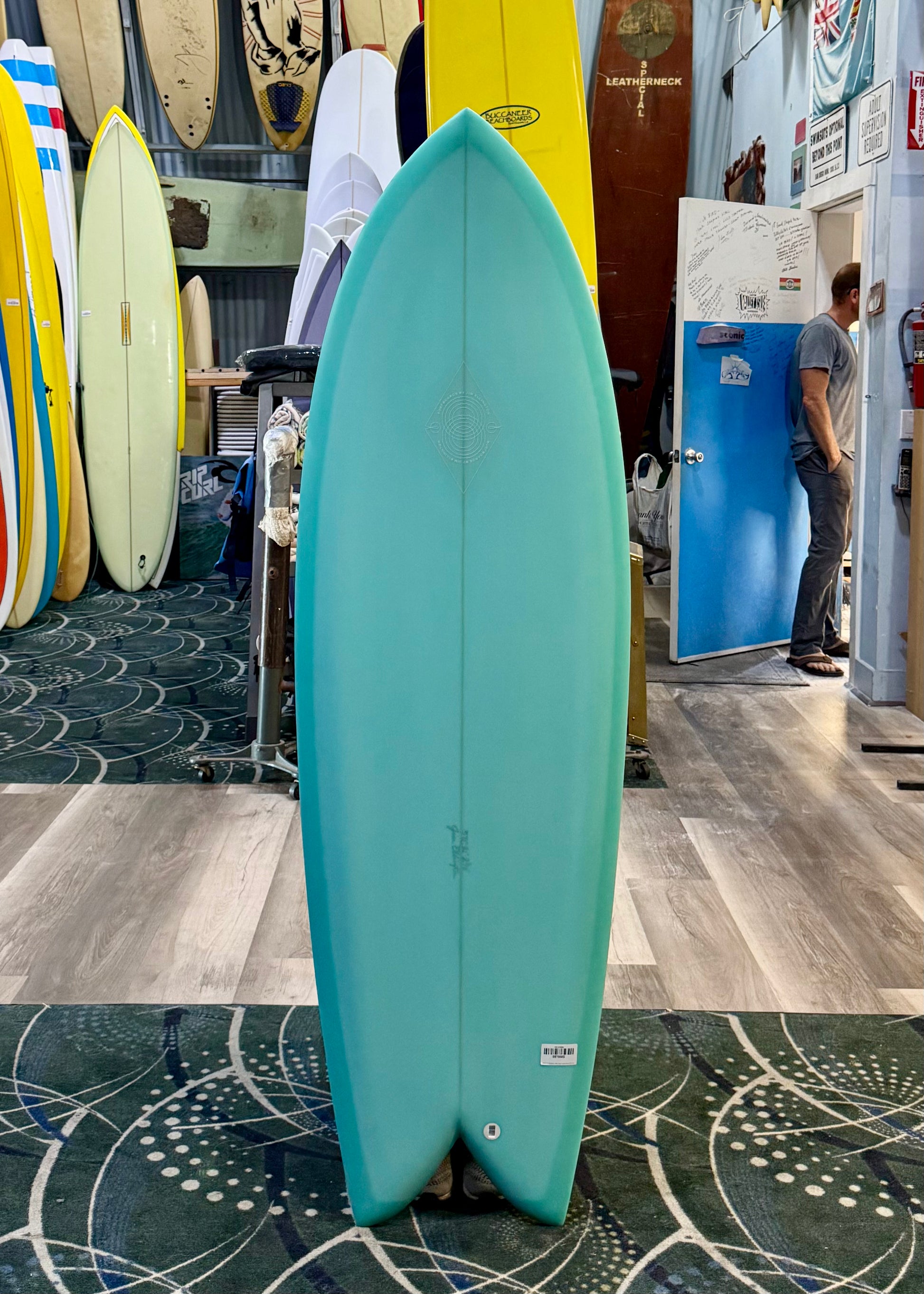 Teal surfboard leaning against a wall in a store with other surfboards and people in the background.