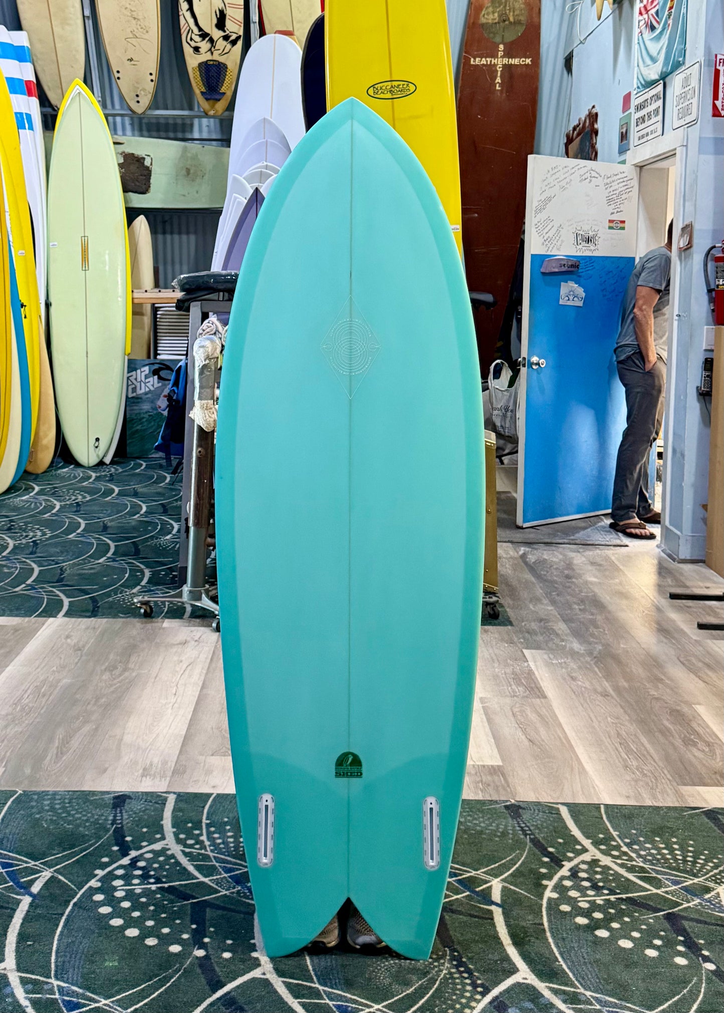 Turquoise surfboard in a store with other surfboards and a person in the background