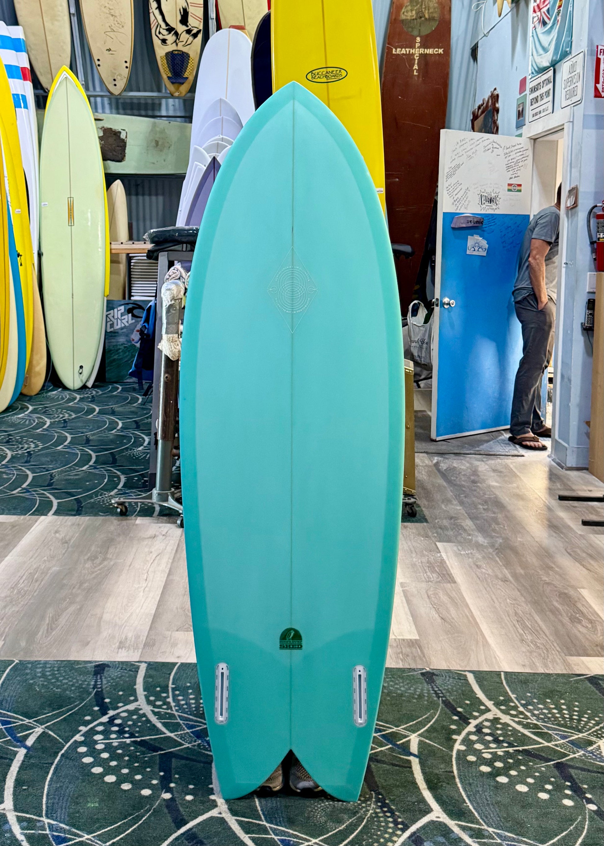 Turquoise surfboard in a store with other surfboards and a person in the background