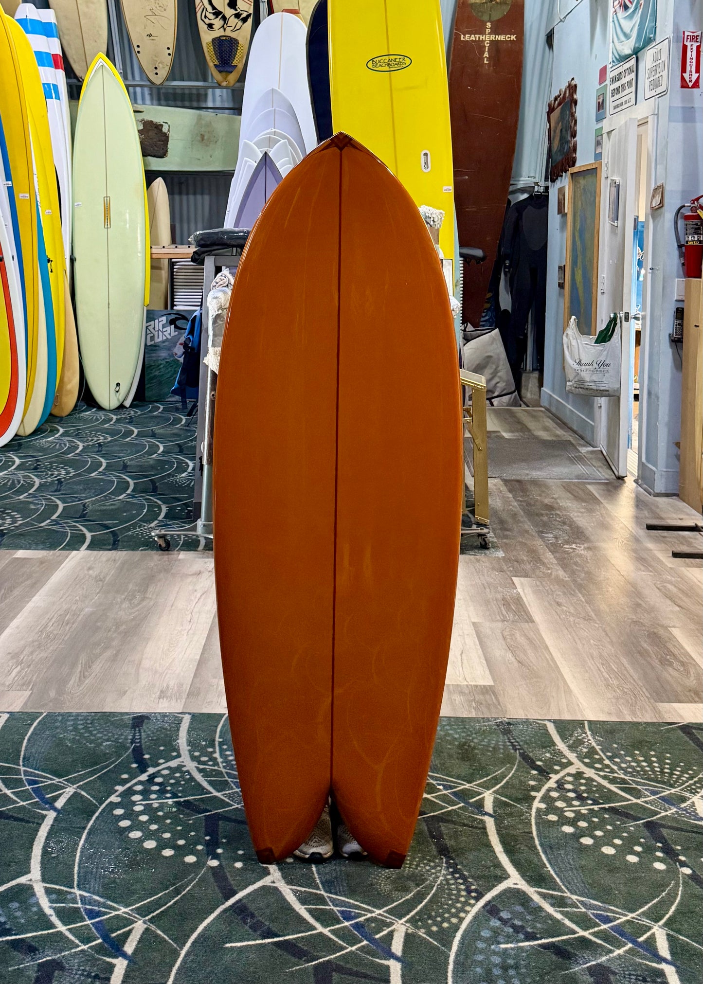 Orange surfboard standing upright on a patterned floor with other surfboards in the background.
