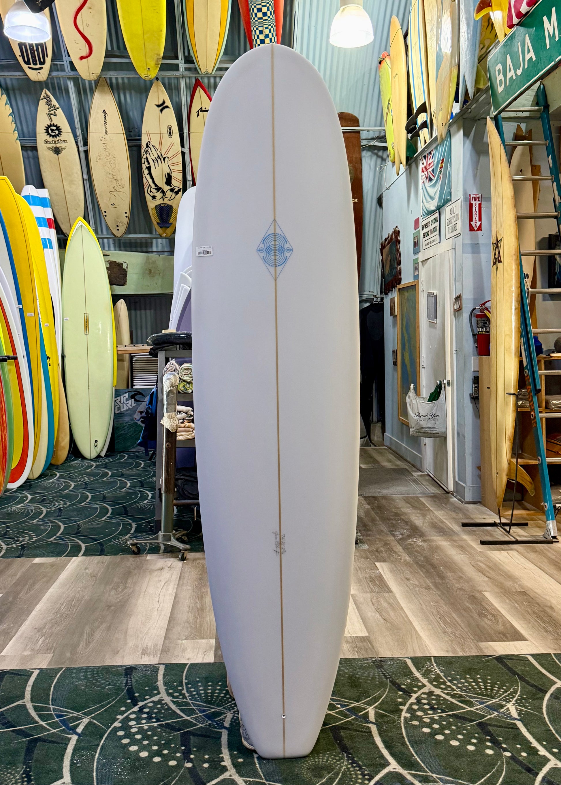 White surfboard with a blue logo in a store with various surfboards and signs on the wall.
