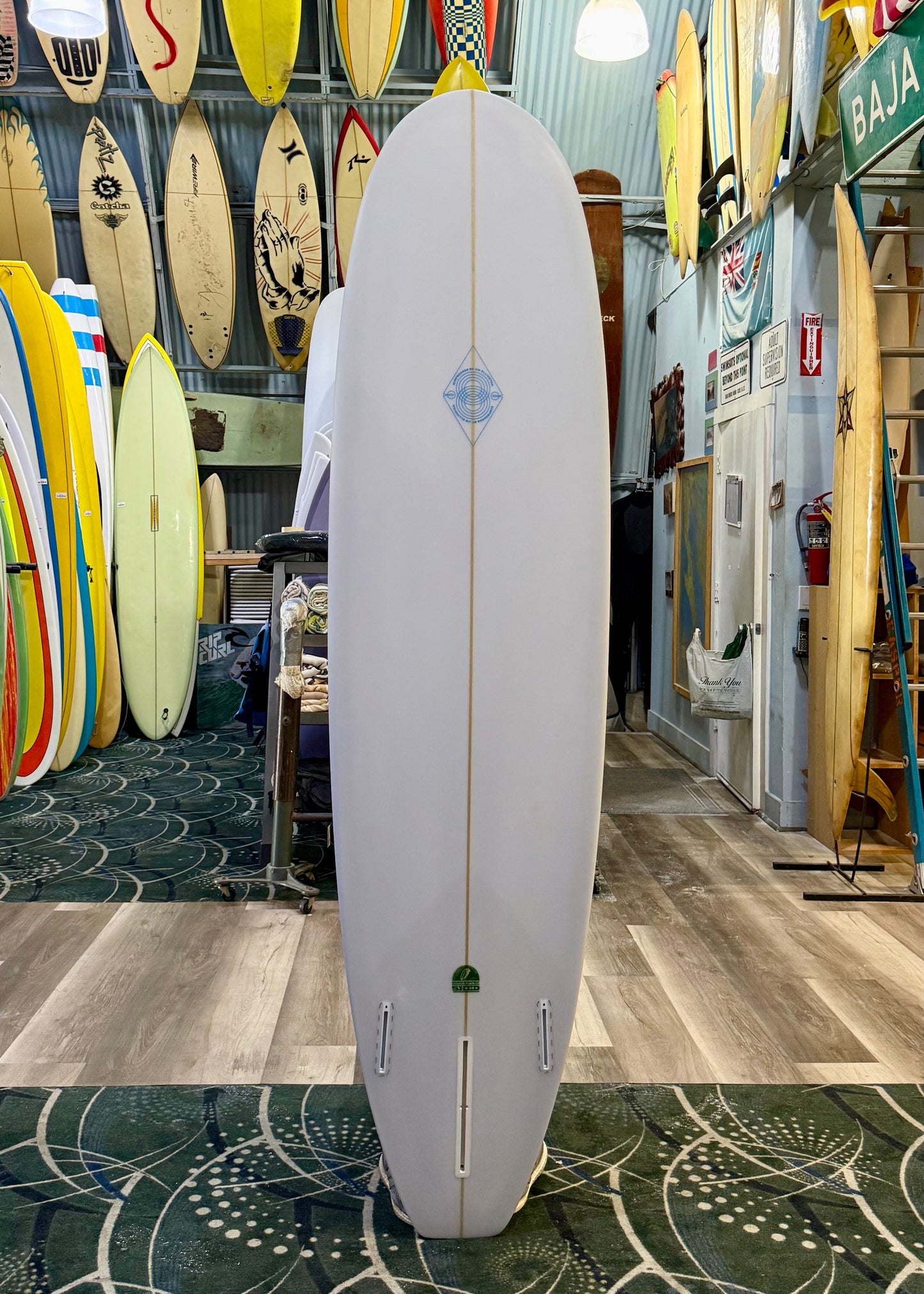 White surfboard with a blue logo in a surf shop with various surfboards on the wall.